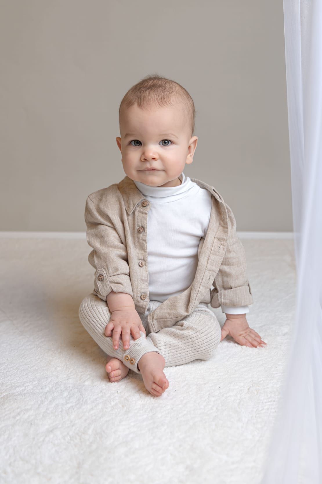 Baby in Leinenhemd sitzt auf weißem Teppich und schaut in die Kamera